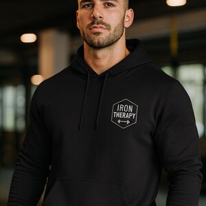 May include: Black hoodie with a front pocket and drawstring hood. The chest features a white logo that reads "IRON THERAPY" above a barbell graphic. The hoodie is worn by a person in a gym setting.