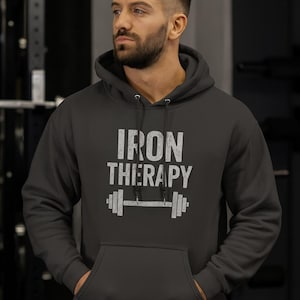 Może przedstawiać: Ciemnoszara bluza z kapturem z białym napisem "IRON THERAPY" i grafiką sztangi. Bluza ma przednią kieszeń i kaptur. Osoba na zdjęciu ma na sobie bluzę.