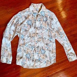 May include: A long-sleeved button-down shirt with a nature-themed pattern. The shirt features a repeating design of deer, trees, and flowers in shades of beige, blue, and brown. The shirt has a classic collar and buttoned cuffs.