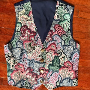 May include: A vintage vest with a colorful floral pattern in shades of pink, green, and blue. The vest has a navy blue back and lining, with gold-colored buttons down the front. The vest is a classic style, perfect for layering.