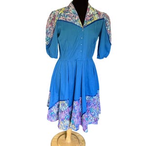 Può includere: Un vestito blu vintage con colletto e maniche a fantasia floreale. L'abito presenta un'abbottonatura frontale e una gonna svasata con orlo a fantasia floreale. L'abito è esposto su un manichino di legno.