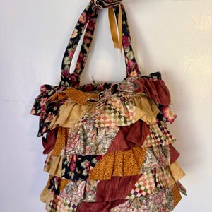 May include: A handmade tote bag with a ruffled design. The bag features a patchwork of floral and patterned fabrics in shades of burgundy, gold, and cream. The handles are made of a floral fabric with a dark background. The bag is hanging against a white wall.