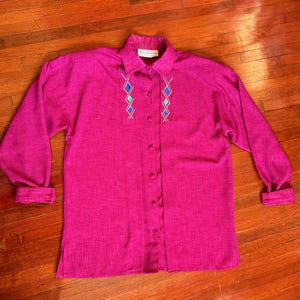 May include: A magenta button-down shirt with long sleeves. The shirt features a collared neckline and embroidered geometric designs in blue and green. The fabric has a textured appearance. The shirt is laying on a wooden surface.