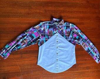 May include: A long-sleeved button-down shirt with a light blue denim-like front and a patterned back and sleeves. The pattern includes purple, teal, brown, and white geometric shapes. The shirt has a classic collar and buttoned cuffs.