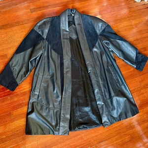 May include: A black leather coat with suede detailing. The coat has a long, flowing design with a shawl collar and pockets. The sleeves and upper chest have black suede panels. The coat is displayed on a wooden floor.