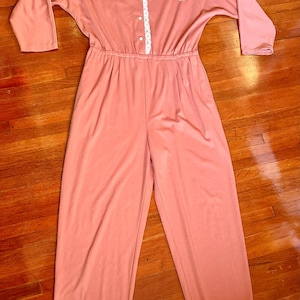 May include: A coral-colored jumpsuit with long sleeves and a button-up front. The shoulders are adorned with floral and lace embellishments. The waist is cinched with an elastic band, and the pants are wide-legged.