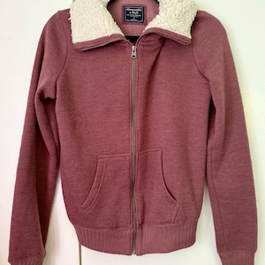May include: A maroon zip-up jacket with a cream-colored, fuzzy collar. The jacket has a ribbed texture, a full zipper, and two front pockets. The brand tag is visible on the collar. A cozy outerwear item.