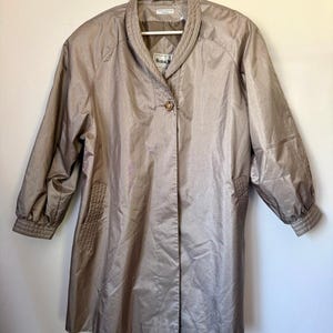May include: A tan-colored, long-sleeved coat with a button closure and a wide collar. The coat has a slightly shiny appearance and a tailored fit. The sleeves are gathered at the cuffs. The coat is hanging on a white hanger.