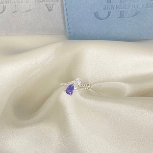 May include: A silver ring with a purple gemstone and a small clear stone. The ring is on a white fabric background.