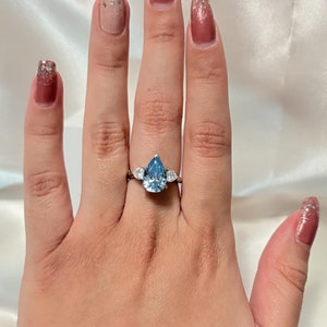 May include: A hand wearing a silver ring with a large pear-shaped blue gemstone and two smaller round gemstones. The hand is also wearing a silver bracelet with small round gemstones. The nails are painted a light brown color with silver glitter.