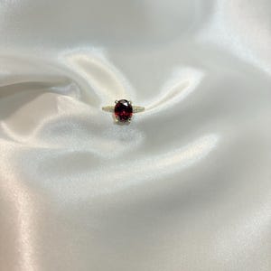 May include: A gold ring with a large, oval-cut, deep red gemstone as the centerpiece. The band is thin and appears to be adorned with small, clear accent stones. The ring is set against a backdrop of white, silky fabric.