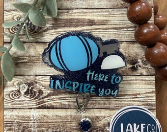 May include: A badge reel featuring a dark blue glitter background with a light blue graphic of a medical bulb syringe and the text Here to INSPIRE you. A circular sticker with the text LAKECO CUSTOMS is visible in the bottom right corner.
