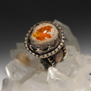 Op de afbeelding: Een zilveren ring met een grote, ronde, oranje opaal-steen gezet in een getextureerde zilveren bezel. De bezel is versierd met een rij kleine sterren.