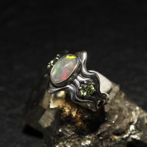 May include: A silver ring featuring an oval opal gemstone with iridescent colors, flanked by two smaller green gemstones. The ring has a unique, flowing design, set against a dark background.