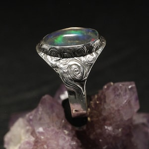 Puede incluir: Un anillo de plata con una piedra preciosa de ópalo ovalada e iridiscente. La banda y el engaste del anillo presentan un patrón detallado y arremolinado. El ópalo muestra tonos verdes, azules y rojos. El anillo se exhibe sobre un cristal púrpura.