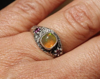 May include: A silver ring with an oval, iridescent opal center stone. The ring has a textured band and is accented with small, round, pink gemstones. The opal displays shades of yellow, orange, and green.