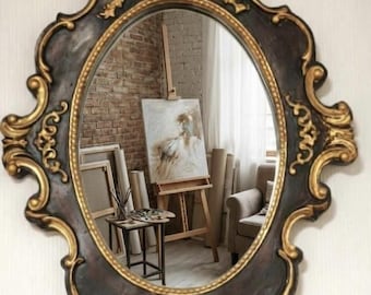 Puede incluir: Un espejo de pared ovalado con un marco marrón oscuro y detalles de volutas pintadas en oro. La superficie de cristal reflectante muestra una habitación con un caballete de artista con un lienzo, una mesa auxiliar y una silla.