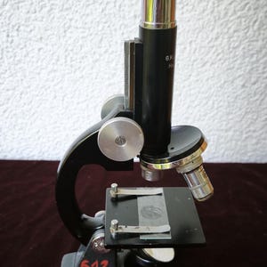 May include: A black metal compound microscope featuring a silver eyepiece tube, a square stage with two metal clips, and a coarse adjustment knob. The base is marked with the red painted number 612, and the body displays the text G.K. and H.