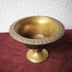 May include: A brass pedestal bowl featuring a wide, flared rim decorated with a repeating oval pattern. The interior of the bowl shows a darkened, aged patina, while the exterior base is smooth and circular, supporting the fluted body of the vessel.