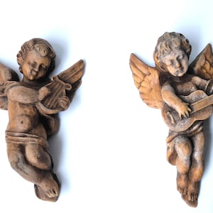 Puede incluir: Dos esculturas de querubines de estilo antiguo. Uno toca el arpa, el otro la guitarra. Ambos tienen un tono marrón cálido con alas y rasgos faciales detallados. Estas figuras decorativas probablemente están hechas de un material compuesto.
