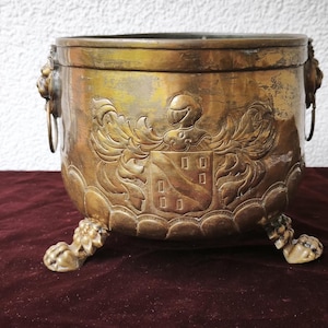 Könnte beinhalten: Antiker Messing-Pflanztopf mit detailliertem Wappen und Löwenkopfgriffen. Der Pflanztopf steht auf drei Löwenpfotenfüßen. Das gealterte Messing hat einen warmen, goldenen Farbton, der zu seinem Vintage-Appeal beiträgt.