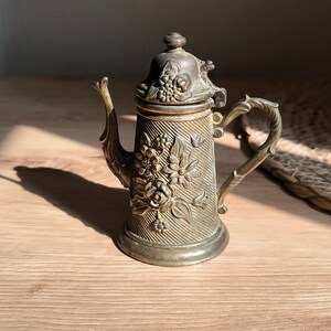 Könnte beinhalten: Eine antike, goldfarbene Teekanne aus Metall mit floralem Design. Die Teekanne hat einen Deckel mit Knauf und einen Ausguss. Die Teekanne steht auf einer Holzoberfläche.