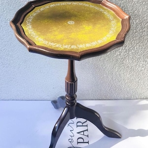Puede incluir: Una pequeña mesa auxiliar de estilo antiguo con borde festoneado. La mesa presenta un marco de madera marrón oscuro y una tapa amarilla con un borde blanco decorativo. La mesa mide aproximadamente 71 cm de altura.