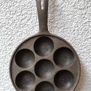 Può includere: Padella in ghisa marrone scuro con sette incavi circolari per la cottura. La padella ha un lungo manico con un foro per appenderla. È appoggiata su una superficie di legno contro un muro bianco strutturato.