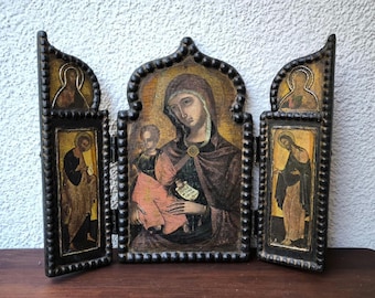 May include: A triptych icon featuring religious figures. The central panel depicts a woman holding a child, with two side panels showing other figures. The artwork is framed in a dark, ornate wooden frame with a beaded border.