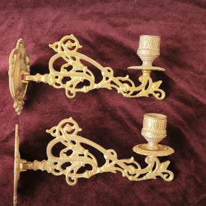 May include: Two ornate, gold-colored wall sconces. Each sconce features a decorative backplate, an elaborate scrolling arm, and a candle cup. The sconces are made of metal and are designed to hold candles, adding a touch of elegance to any room.