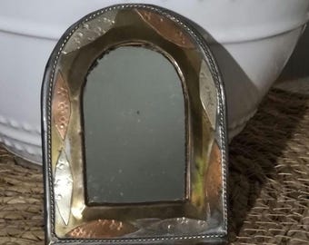 May include: A small, arched, silver-toned mirror with a decorative frame made of copper, brass, and silver. The frame has a geometric pattern with a diamond shape in the centre.