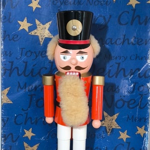 Op de afbeelding: Een traditioneel houten notenkrakerfiguur met een rood-zwart uniform, een witte baard en een groene basis. De notenkraker staat voor een blauwe achtergrond met gouden sterren en de tekst "Joyeux Noel" en "Christmas".