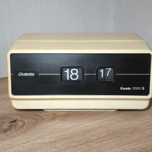 May include: A white flip clock with black numbers displaying 18:17. The clock has a black face with the word "Dielente" and "Kundo 2000 S" printed on it.