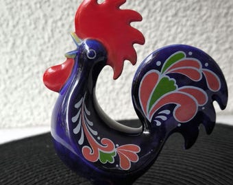 May include: A ceramic rooster figurine in shades of blue, red, and green. The rooster has a vibrant red comb and wattle, with a detailed tail and wing design. The base is round, and the figurine is set against a neutral background.