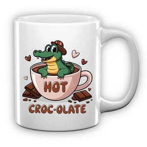 Pode incluir: Caneca de cerâmica branca com um jacaré de desenho animado numa chávena rosa de chocolate quente. A caneca tem as palavras "HOT CROC-OLATE" impressas, juntamente com quadrados de chocolate e corações.