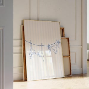 May include: A framed artwork featuring a blue line drawing of a bikini hanging on a clothesline. The artwork has a gold frame and is leaning against a wall with other framed pieces. The background is a light-colored wall with vertical stripes.