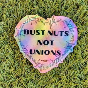 May include: A holographic sticker in the shape of a heart with barbed wire around the edge. The sticker says "Bust Nuts Not Unions" in black text.