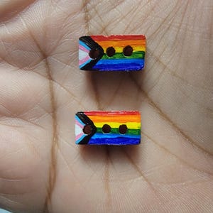 May include: Two rectangular wooden buttons, each painted with a rainbow gradient and the transgender pride flag. The buttons have three holes for sewing and are held in a hand.