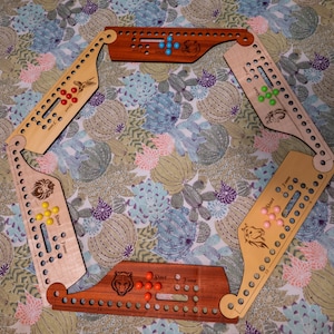 Può includere: Gioco da tavolo in legno con sei sezioni disposte a esagono. Ogni sezione presenta un diverso design di animali e pioli colorati. Il gioco è ambientato su uno sfondo di tessuto a fantasia floreale.