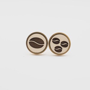 May include: Two round wooden earrings with a coffee theme. One earring features a single coffee bean, while the other has three. The earrings have a light beige background with a dark brown coffee bean design and a gold-colored border.