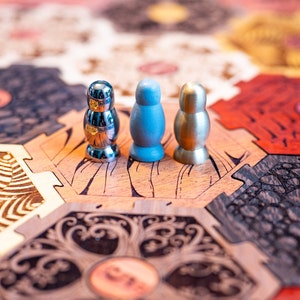 Puede incluir: Tres piezas de juego en un tablero de juego de madera. Las piezas son plateadas, azules y doradas. El tablero está hecho de madera y tiene un patrón hexagonal.