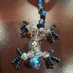 May include: A beaded lizard figurine with a long tail and four legs. The lizard is made of clear, blue, and black beads. The body is a mix of faceted and round beads. The tail is a string of blue and black beads.