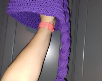 May include: A handmade, purple crochet hat with a long, braided tail. The hat is a vibrant shade of purple and appears to be made of soft yarn. The braid ends in a tassel. The hat is held up by a person's arm.