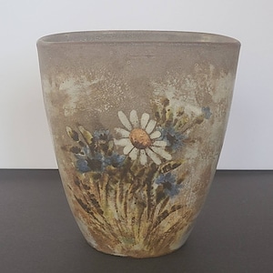 May include: A ceramic vase with a slightly textured, light brown exterior. The vase features a painted floral design with a large white daisy, blue flowers, and green foliage. The vase has a rectangular shape with rounded corners.