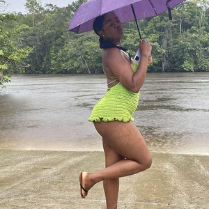 May include: A woman wearing a green crochet swimsuit and black sandals stands in the rain under a purple umbrella. She is standing on a sandy beach with a river in the background.