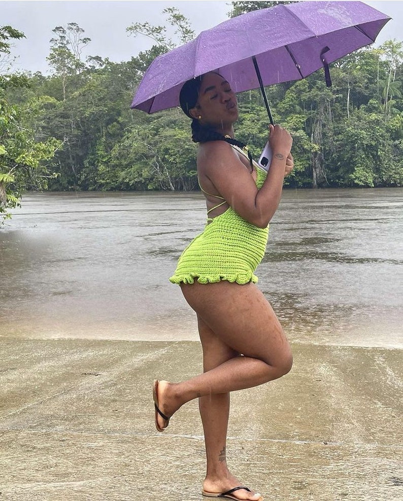 May include: A woman wearing a green crochet swimsuit and black sandals stands in the rain under a purple umbrella. She is standing on a sandy beach with a river in the background.