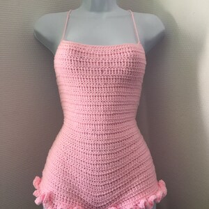 May include: A pink crocheted bodysuit with a ruffled hem. The bodysuit has thin straps and a fitted silhouette.