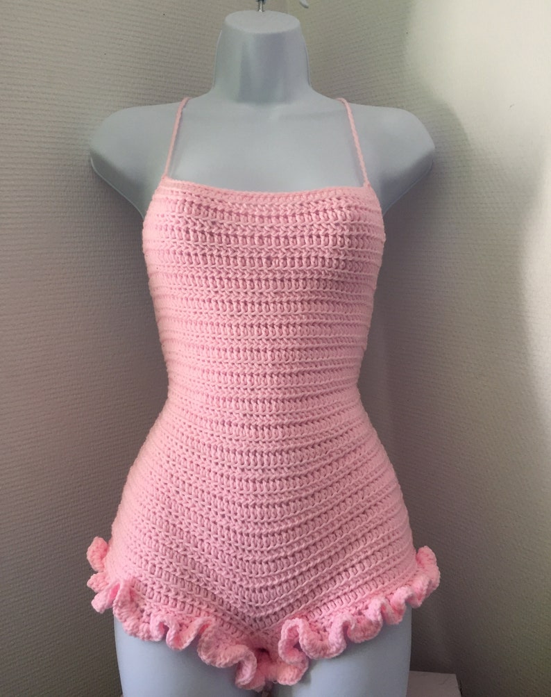 May include: A pink crocheted bodysuit with a ruffled hem. The bodysuit has thin straps and a fitted silhouette.