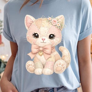 May include: Light blue t-shirt featuring a patchwork kitten design. The kitten has a pink bow, a floral crown, and a heart-shaped pendant. The design incorporates soft pink, peach, and cream colors, creating a whimsical and charming aesthetic.
