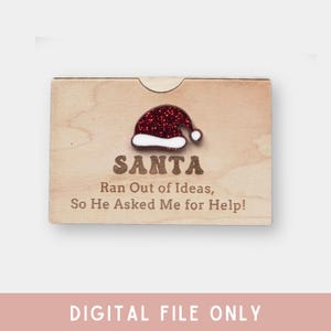 Pu&ograve; includere: Una scatola regalo in legno con un cappello di Babbo Natale rosso glitterato in cima. La scatola ha il testo "SANTA Ran Out of Ideas, So He Asked Me for Help!" stampato su di essa.